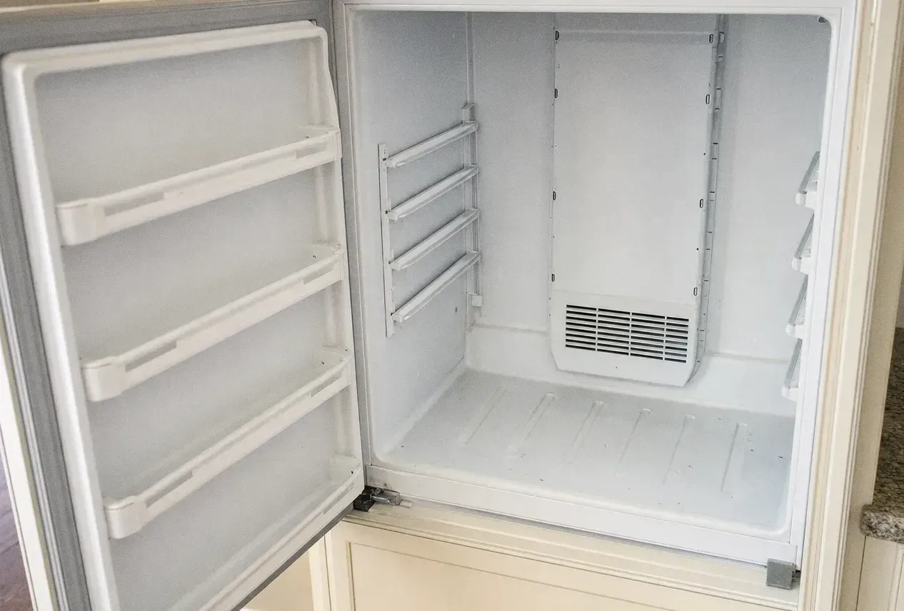 The empty interior of a refrigerator with the door open, shelf rails on the side walls and a vent grille on the rear panel